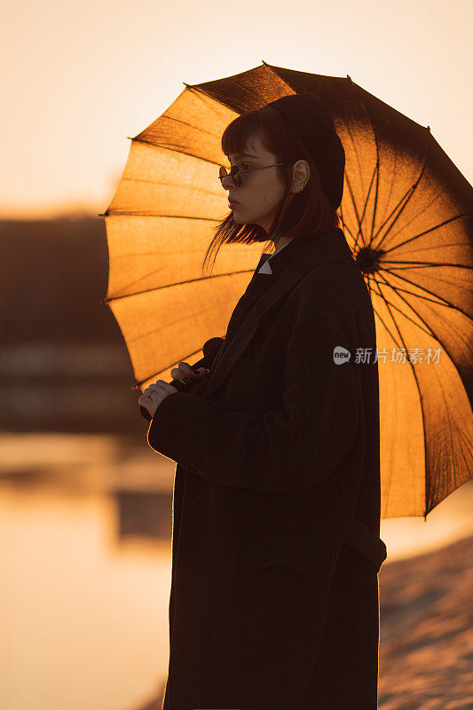 红发女孩在日落背景下的户外。哥特式风格。亚洲时尚模特，黑色外套，雨伞，自然。黑色的太阳镜。时尚的衣服。青年现代风格。亚文化