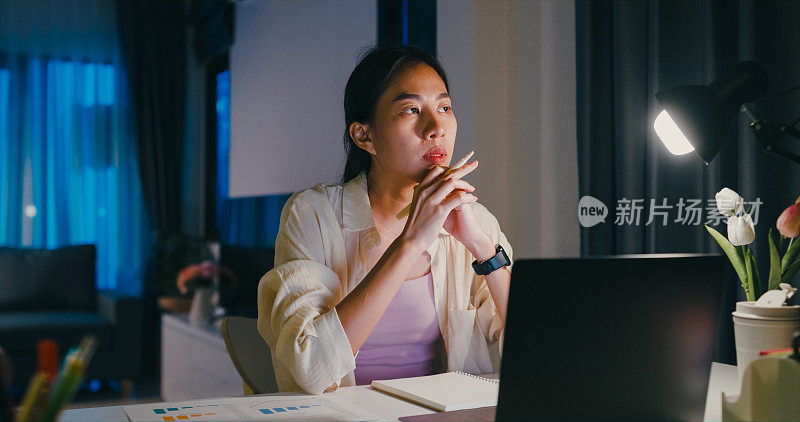 年轻的亚洲女性穿着正装衬衫坐在桌子前与笔记本电脑和文书工作想主意使紧急报告会议加班到晚上在家。