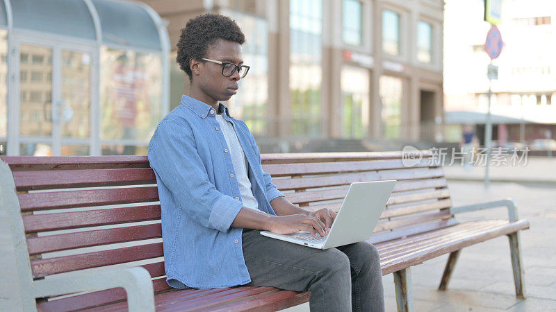 忙碌的非洲青年坐在户外长凳上使用笔记本电脑