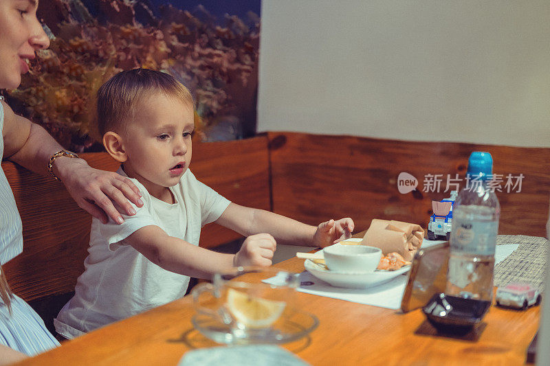 快乐的年轻怀孕妈妈和蹒跚学步的儿子正在等待晚餐，同时在他们的手机上看视频