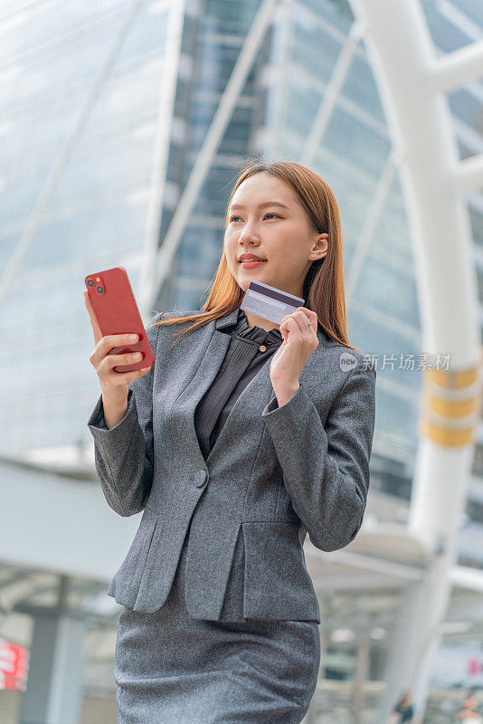 一名年轻的亚洲女商人穿着灰色的办公套装，在商业区用她的智能手机使用信用卡进行在线交易，她微笑着自信