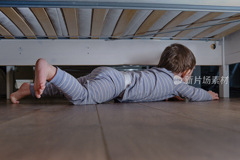 蹒跚学步的婴儿爬着藏在床底下。孩子爬到沙发下面，坐在地板上。孩子男孩年龄一岁八个月，全长
