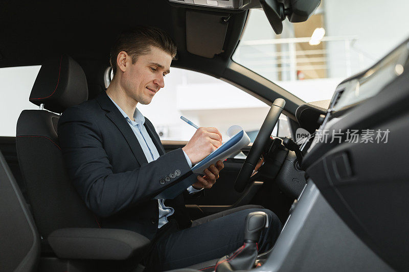 积极的年轻汽车销售员坐在新车里，做检查，在经销商试驾期间做笔记