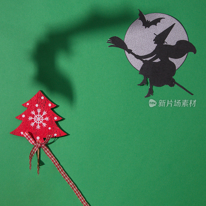 一个女巫骑着扫帚飞到月亮旁边，带着一只蝙蝠和一只蝙蝠的影子飞过圣诞树，在绿色的背景上。复制空间和最小的平面场景。