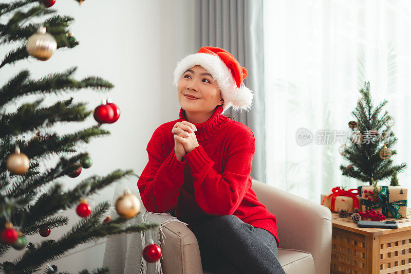 亚洲妇女开心地在圣诞节那天许愿，坐在装饰着圣诞树的客厅里。