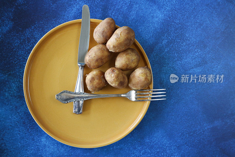 黄色盘子用不锈钢刀叉形成钟面和指针，小块堆土豆，蓝色背景，高架视野，复制空间，饮食和间歇性禁食的概念