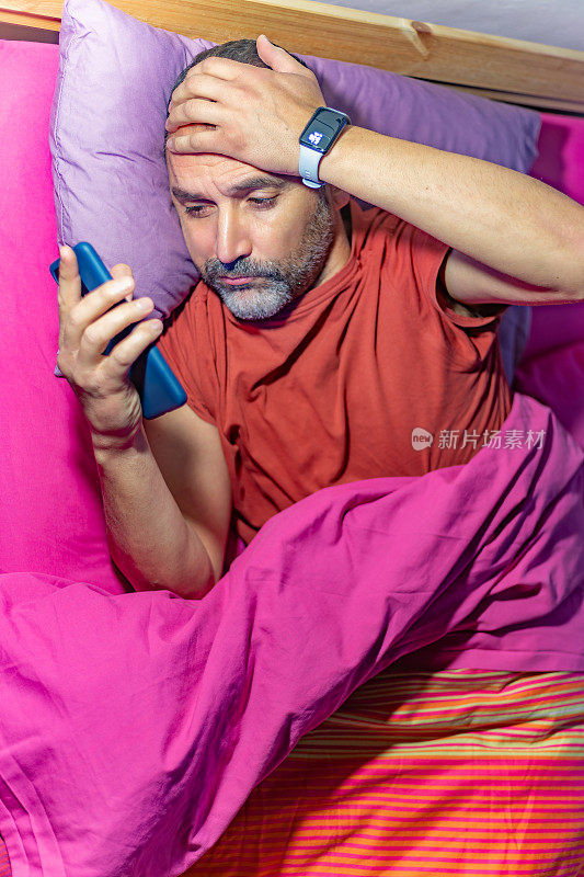 俯视图:一个长着胡子的成熟男子躺在床上，一边看手机，一边把一只手放在额头上，做出不舒服的手势。