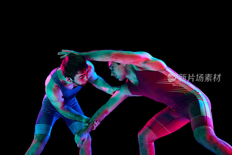 年轻的熟练的摔跤手在蓝色和红色统一的手在中立的位置在他们的脚在黑色背景混合霓虹灯。