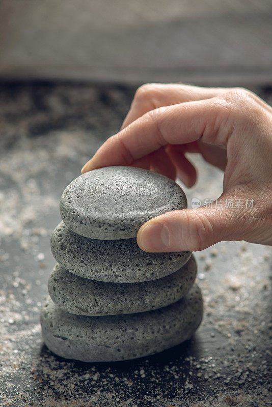 在黑色的背景上，一块块石头堆叠成天然肥皂的形状，上面覆盖着海盐，一只手把石头放在上面。有机化妆品和水疗的概念
