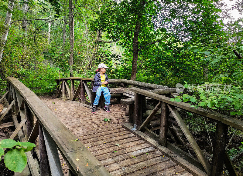 一个8岁的小女孩坐在一座废弃的旧木桥上，桥上有一个休闲区，桥对面是森林里一条狭窄的河流。