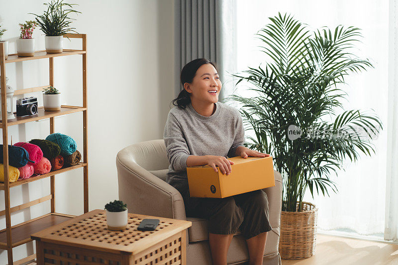 亚洲女人拿着纸盒，开心兴奋地坐在家里客厅的扶手椅上。