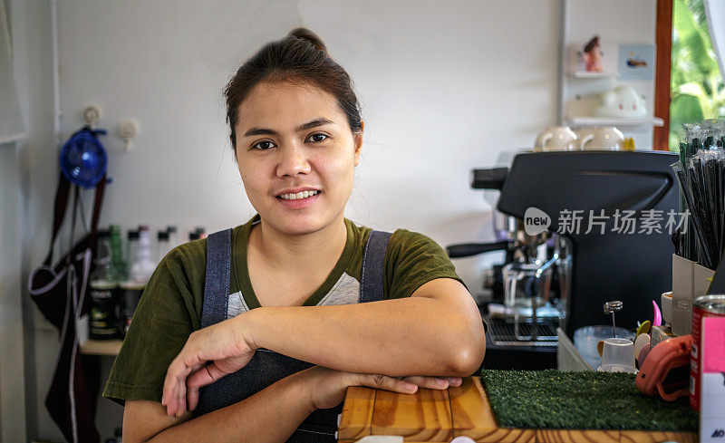 站在柜台后面的亚洲泰国女咖啡店老板的肖像。自信的女咖啡师站在柜台后面。一位穿着围裙的女咖啡馆老板微笑着看着镜头。