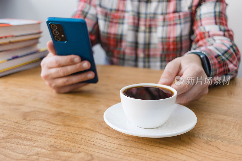 木桌上的一杯咖啡，一个面目全非的人在用智能手机，坐在桌前，手里拿着一摞书，聚焦在前景