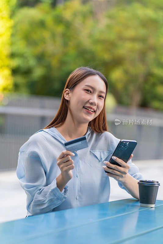 一名亚洲妇女在户外公园用手机进行数字银行信用卡交易，桌上放着一杯咖啡