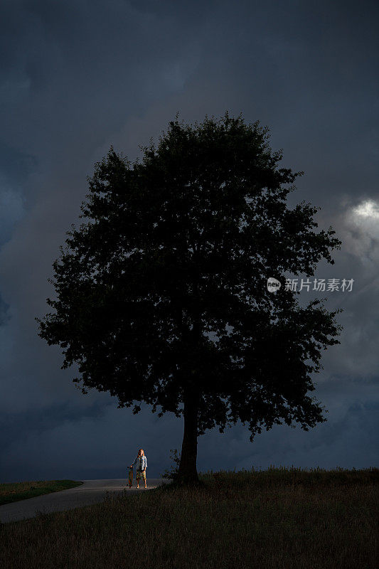 一个小女孩站在路边，手里拿着她的长板，旁边只有一棵树
