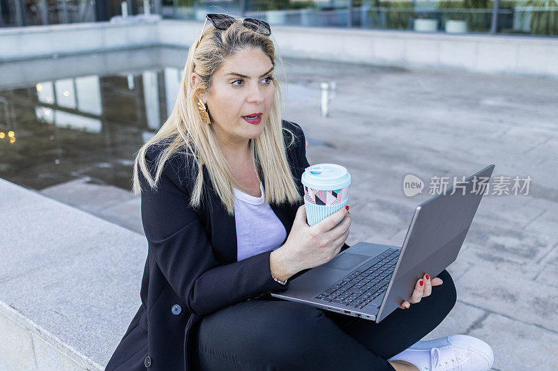 穿着黑色西装的女商人，坐在公司大楼前，用笔记本电脑工作。在户外灵活工作。现代虚拟理财咨询。
