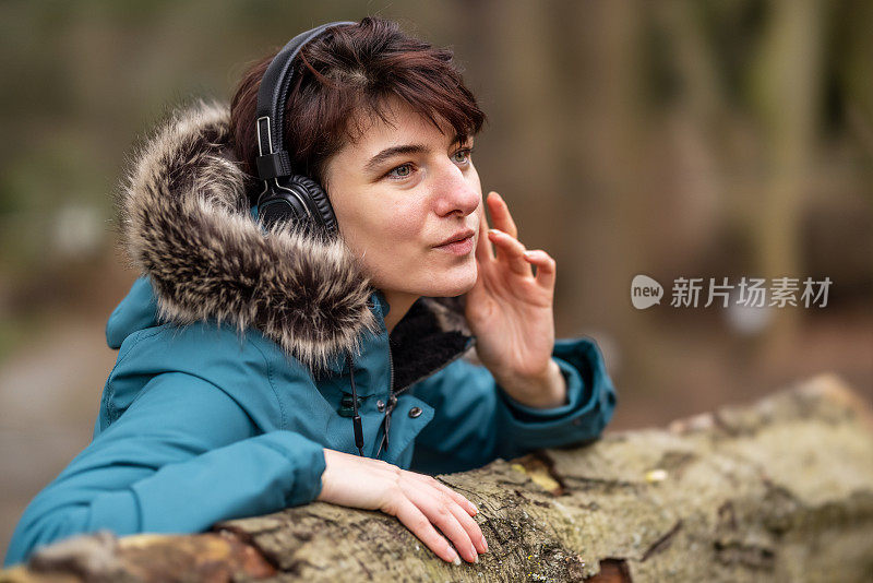 一位穿着蓝色雨衣的年轻女子正戴着耳机坐在户外公园的长椅上听着音乐