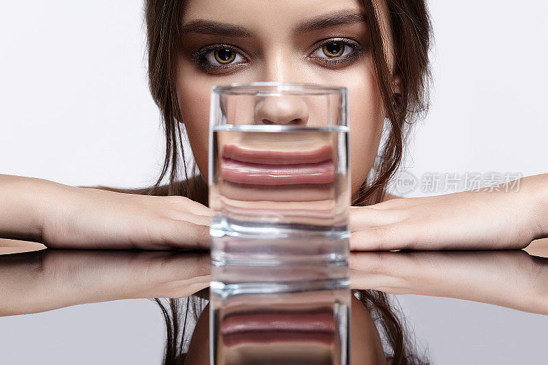 女孩把脸藏在盛有水的杯子后面。镜桌上年轻女子的光学扭曲肖像。