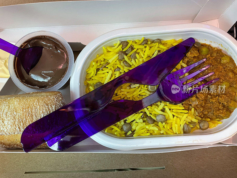 一次性的特写图像，纸板飞机食物托盘，内含印度咖喱饭和藏红花饭，面包卷，一次性使用，塑料碗的巧克力慕斯，一次性塑料紫色餐具，提高的看法