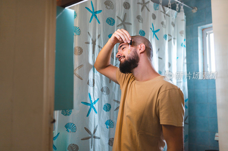 一个秃顶的年轻人对着浴室的镜子直接在胡子上涂胡子油