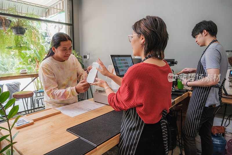 年轻的亚洲女性咖啡师或咖啡店老板在咖啡馆的吧台讨论点饮料菜单并向她的客户推荐咖啡豆