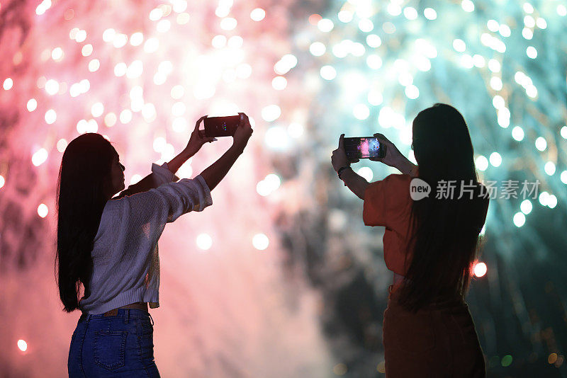 亲密的朋友用智能手机在巨大的烟花庆祝活动中拍照，快乐的年轻女子庆祝除夕