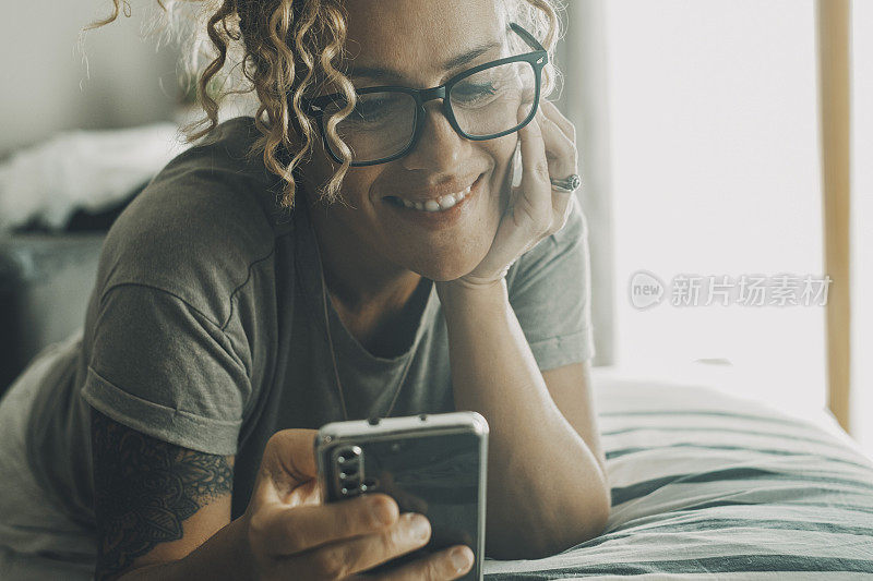欢快快乐的女人躺在床上，用手机聊天和观看在线内容的肖像。拥有智能手机的现代女性。和朋友聊天。放松与科技。写作
