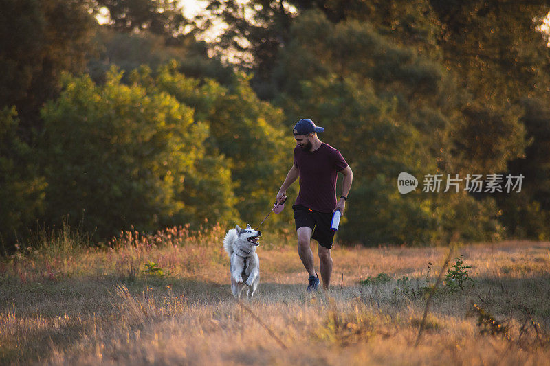 年轻男子与他的哈士奇狗跑在阳光明媚的早晨在大自然