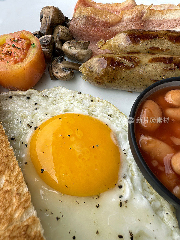 全帧图像的白色盘子包含全英式油炸早餐食材，两根香肠，烤豆小蛋糕，培根片，番茄，蘑菇，煎蛋，吐司片，抬高视图，重点在前景