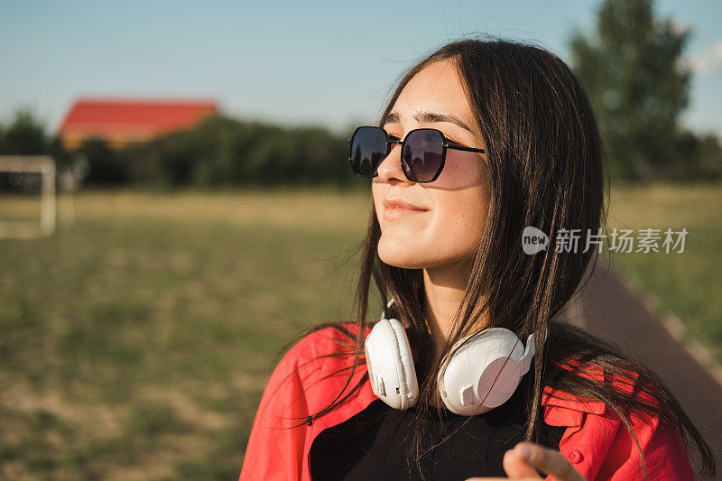 脖子上戴着耳机在户外放松的休闲女性。十几岁的女孩，深色头发，戴着太阳镜，享受着阳光明媚的夏日。