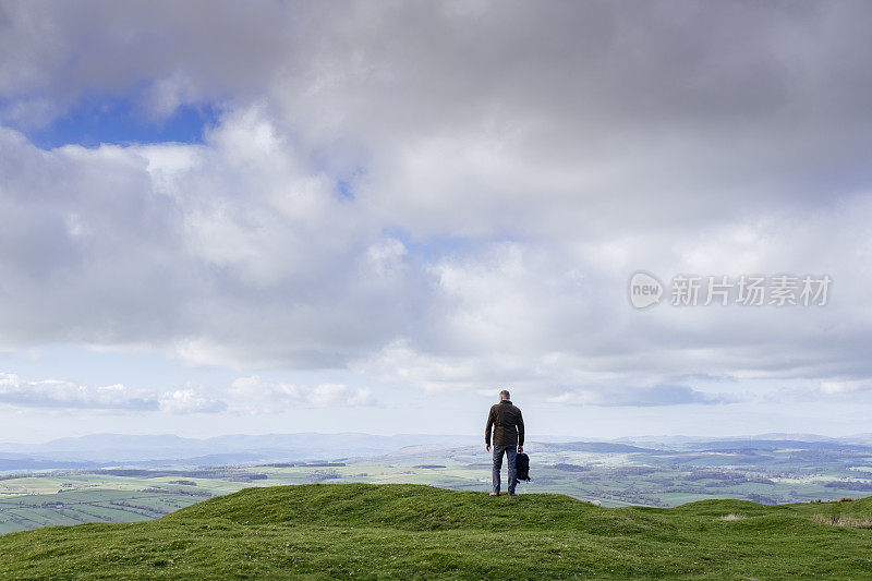 背着双肩包的男子站在山坡上，可以看到广阔的乡村景色。
