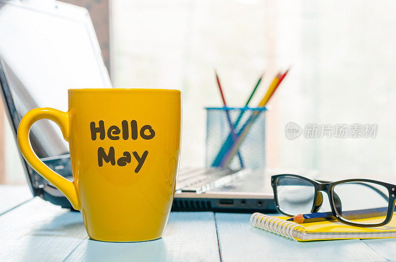你好五月-文字在黄色咖啡杯在商务办公室背景，工作场所与笔记本电脑和眼镜。春天的时候，空白的文字空间