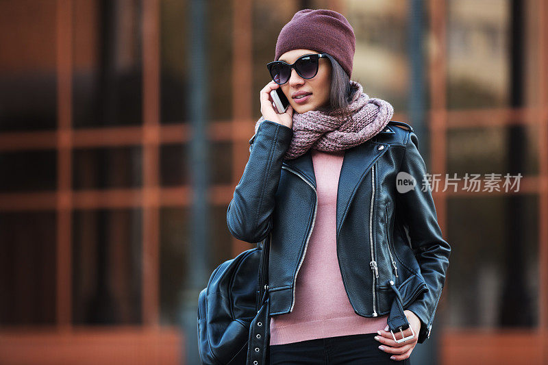 年轻时尚的女人穿着黑色皮夹克在城市街道上使用手机