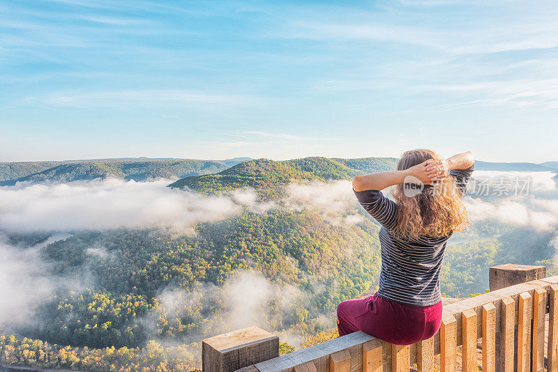 在西弗吉尼亚，一个年轻的女人坐在山的栏杆和浮动的雾云在早晨美景俯瞰，金秋与繁茂的树叶