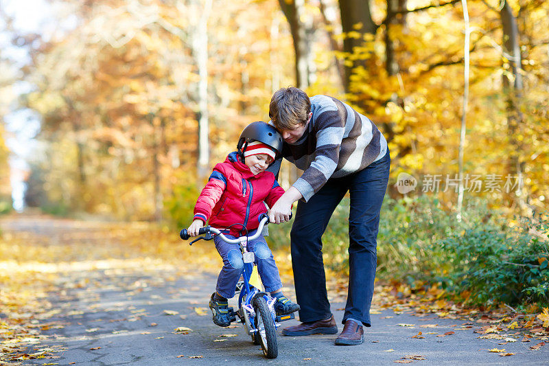 小男孩和他的爸爸在秋天的公园里骑着自行车。爸爸教儿子骑自行车