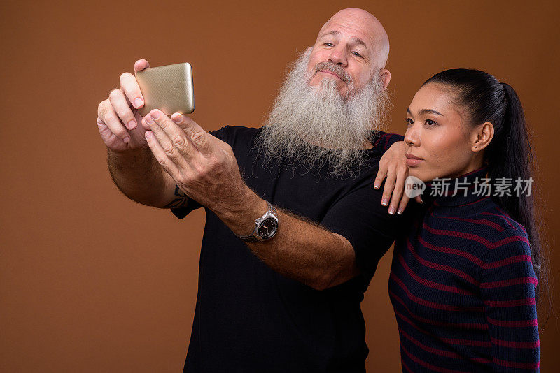 工作室拍摄的成熟的留着长胡子的秃头男人和年轻美丽的亚洲变性女人一起使用手机在有色背景下