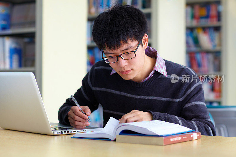 一名亚洲男大学生在图书馆学习，桌上放着笔记本电脑