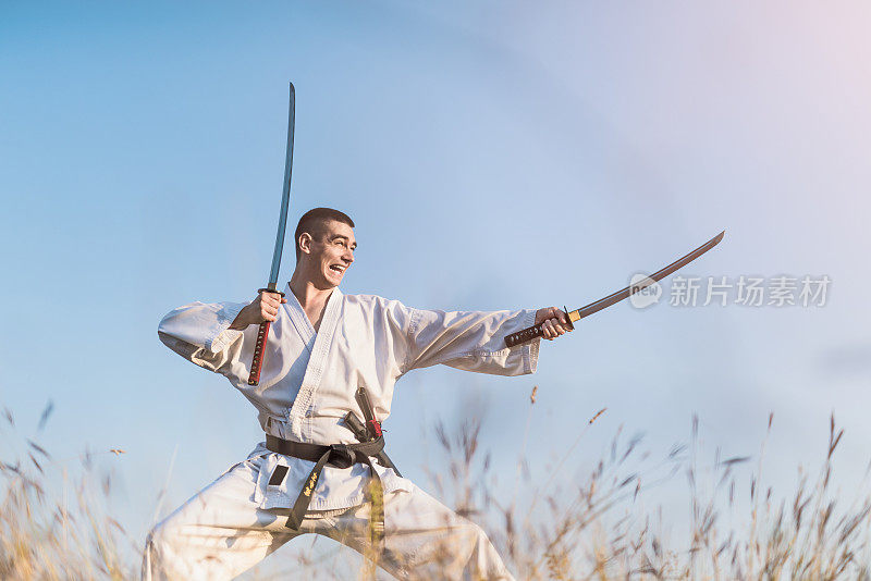 愤怒的年轻武术家练习技巧与两个武士刀在户外的自然。