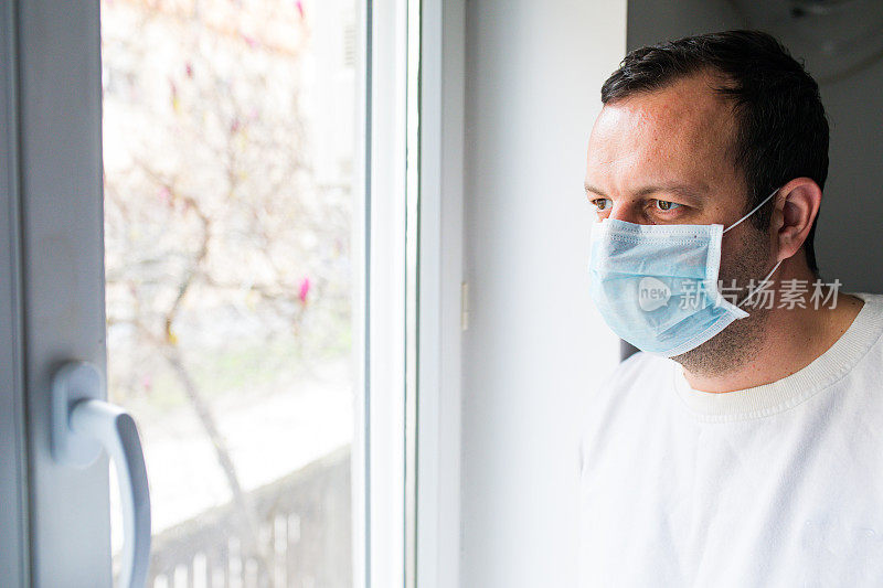 一个戴医用面具的孤独男人透过窗户看。居家隔离自我隔离