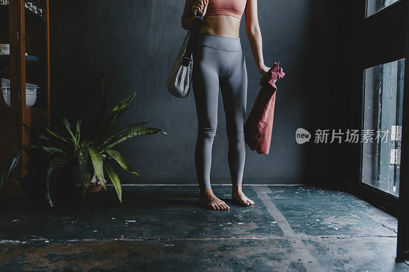 准备在家练瑜伽:一个不知名的年轻女性穿着运动服，包里装着运动垫