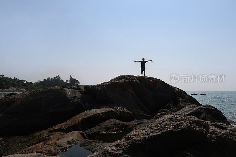 印度男游客假日站在岩石上看以橙色日落为背景的海滩，帕洛伦海滩，印度果阿