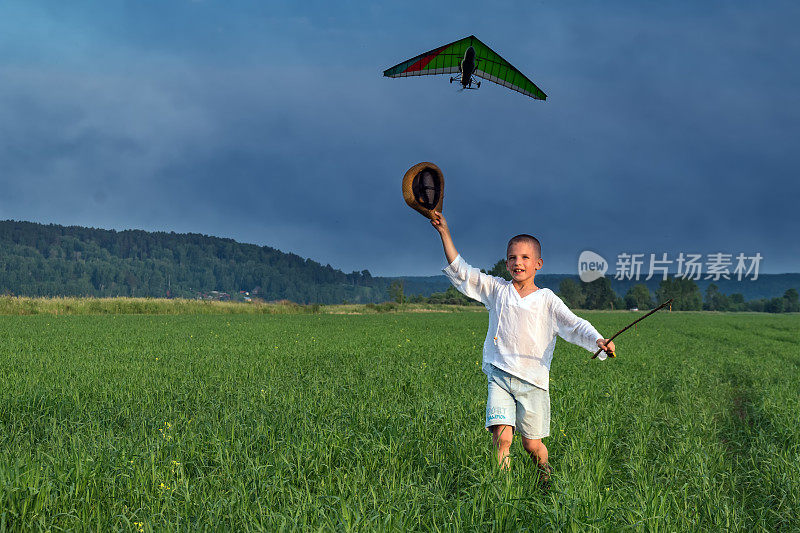 男孩在绿色的田野上挥舞着他的帽子，飞着滑翔伞。男孩挥舞着帽子。