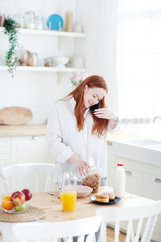 红头发的年轻女子微笑着打电话，在厨房里准备早餐用牛奶做的麦片