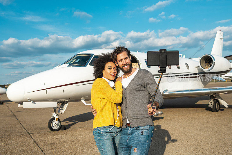 一对著名的年轻夫妇正在一架停在机场跑道上的私人飞机前用装在自拍杆上的智能手机自拍