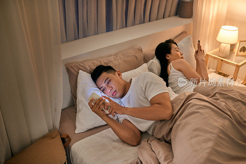一对年轻夫妇躺在床上，背对着对方打电话。