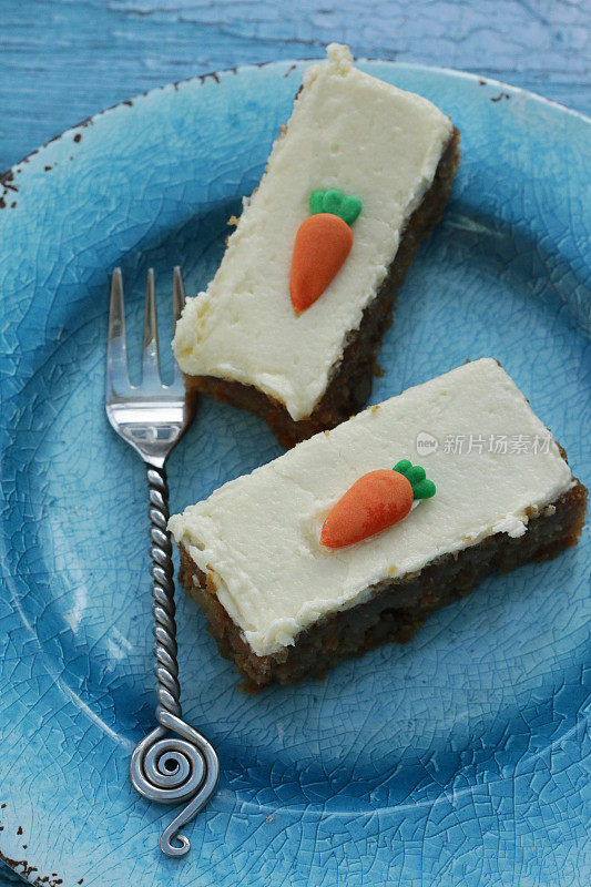 这张照片是自制的两片胡萝卜蛋糕，上面点缀着香草味的奶油，果冻橙片和翻糖胡萝卜糖霜，放在一个用叉子叉着的蓝色盘子里，俯瞰风景