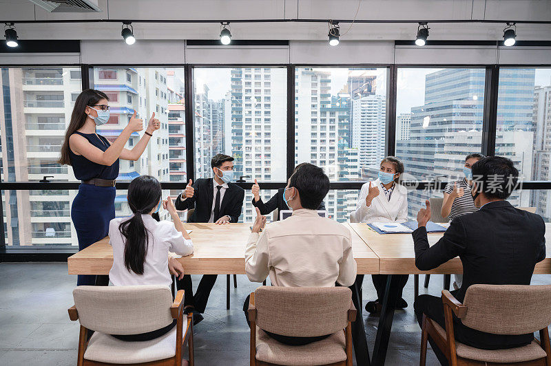 在新常态的办公室里，一群带着口罩的商务人士在会议中竖起大拇指，达成商务协议