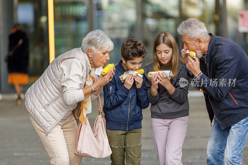 一对老夫妇和孙辈们在城市里散步，享受玉米棒。