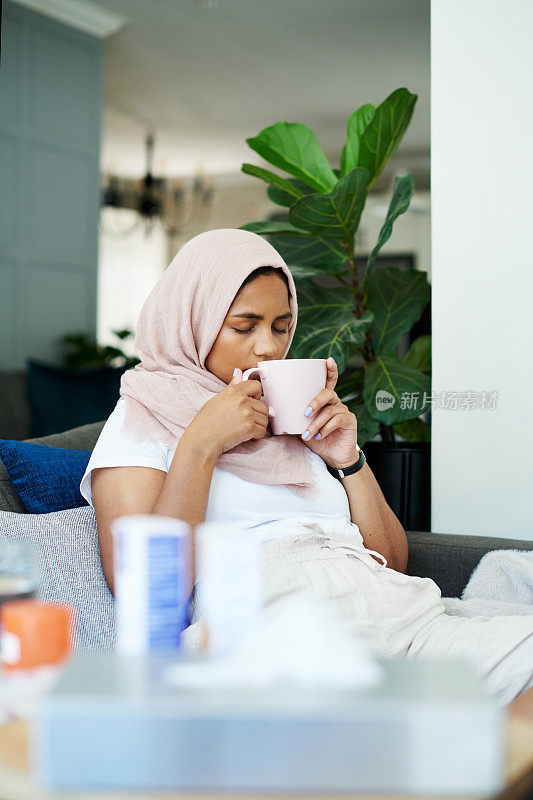 一个迷人的年轻女子独自坐在家里的沙发上，喝着花草茶感觉不舒服
