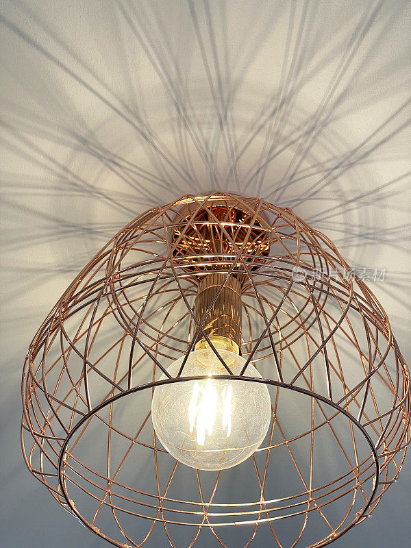 玫瑰金的特写影像，家庭房间天花板上悬挂的铜篮灯罩，由铜棒制成的几何形状灯罩，从下面看